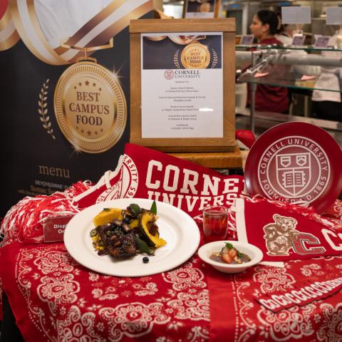 A table with red tablecloth and Cornell pennants, with plates of food and a Best campus Food emblem