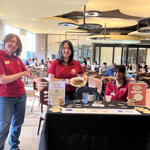 Three people in red Cornell shirts at a table with samples and posters in a dining room, with people eating at tables in the background